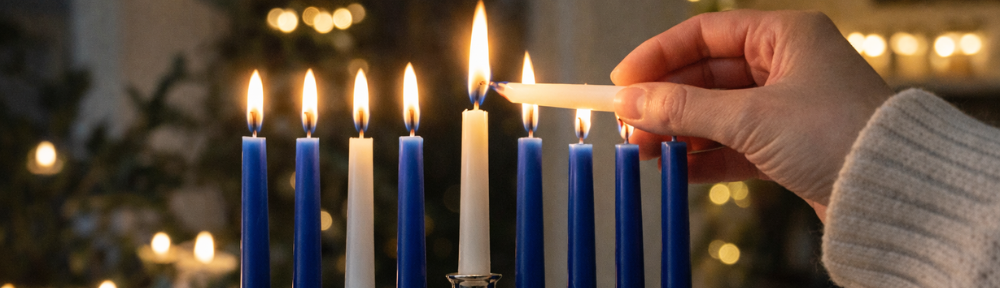 A hand lights a silver menorah with blue and white candles in a softly lit home setting.